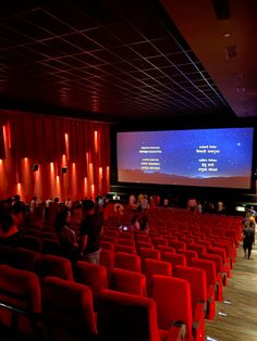 people are standing in an auditorium watching a movie on a large screen with red chairs