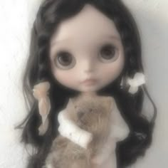 a doll with long black hair holding a brown teddy bear in her arms and looking at the camera