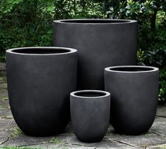 four large pots sitting on top of a stone floor next to green bushes and trees