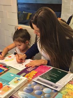 a woman and child are sitting at a table with books on it, while the boy is drawing