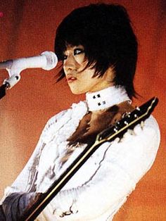 a woman with black hair holding a guitar and singing into a microphone in front of an orange background