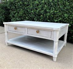 a white coffee table with two drawers in front of a bush and shrub behind it