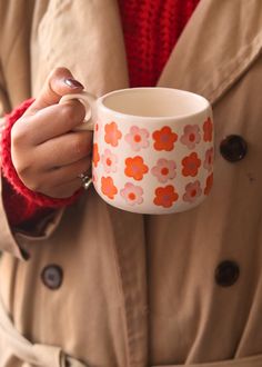 a woman in a trench coat holding a coffee cup with orange flowers painted on it