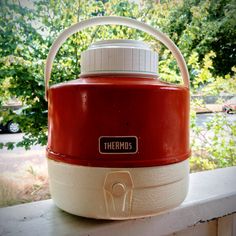 a red and white cooler sitting on top of a window sill next to trees