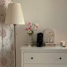 a white dresser topped with a lamp next to a flower filled wallpaper covered wall