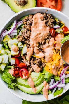 a salad with dressing in a white bowl next to sliced avocado and tomatoes