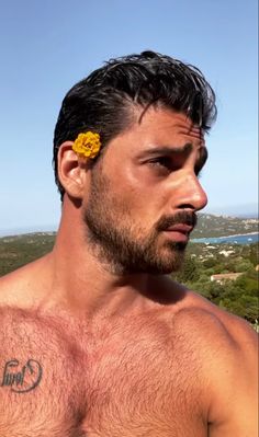 a shirtless man with a yellow flower in his hair looks off into the distance