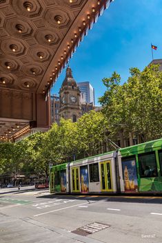 a green and white train traveling past a tall building with a clock on it's face