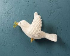 a white knitted bird with yellow feet on a blue wall, holding a flower in it's beak