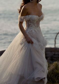 a woman in a wedding dress standing near the ocean with her hand on her hip