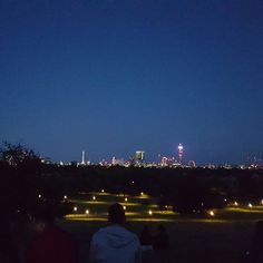 people are sitting on the grass looking at the city lights in the distance, and one person is flying a kite