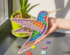 a hand holding a colorful bird shaped wooden puzzle next to a potted plant on a windowsill