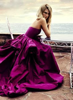 a woman in a long purple dress leaning on a railing near the ocean with her laptop