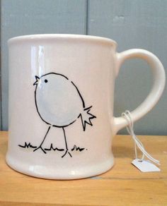 a white coffee mug with a drawing of a bird on it's side sitting on a wooden table