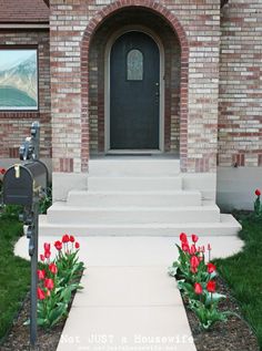 a house with red flowers in front of it and a mailbox on the sidewalk