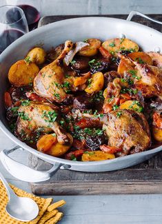 a pan filled with chicken and potatoes on top of a wooden table next to wine glasses