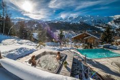 two people sitting in a hot tub on top of a snow covered slope next to a swimming pool