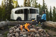 a woman sitting on a log next to a camper and fire in the woods