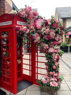 a red phone booth decorated with pink flowers and greenery next to a telephone box