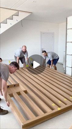 three men working on a wooden floor in a room with white walls and stairs, while another man looks down at the floor