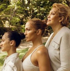 three women sitting next to each other in front of trees and bushes, with one woman looking off into the distance