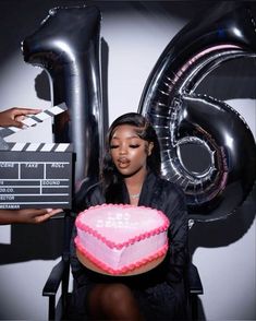 a woman holding a pink heart shaped cake in front of a movie claper and balloons