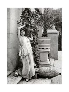 an old photo of a woman leaning against a wall with a vase on her head