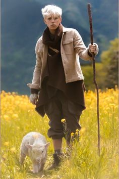 a woman holding a stick and walking next to a small white sheep in a field