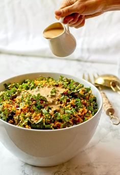 a person pouring dressing into a bowl filled with food on a white tablecloth next to gold utensils