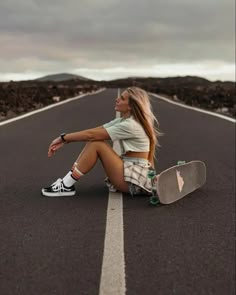 a woman sitting on the side of an empty road with a skateboard in her lap