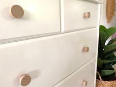 a white dresser with brass knobs in a room next to a potted plant