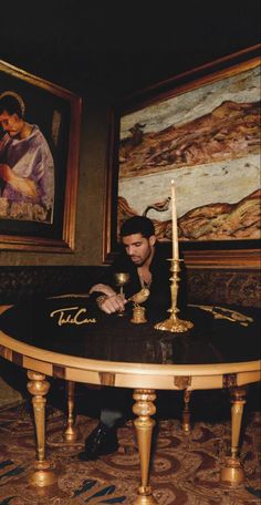 a man sitting at a table with a lit candle in front of him and paintings on the wall behind him