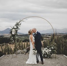 a bride and groom kissing in front of an arch decorated with flowers on top of a mountain