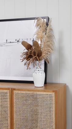 a white vase with some brown flowers on top of a wooden cabinet next to a painting