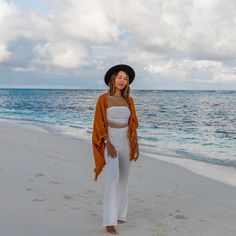 a woman standing on top of a sandy beach next to the ocean wearing a hat