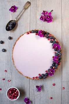 a white cake with purple flowers and berries on it next to a bowl of blueberries