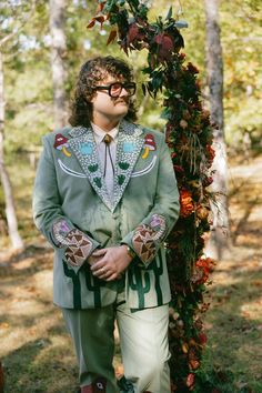 a man in a green suit and tie standing next to a flower covered arch with leaves on it