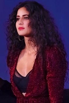 a woman with curly hair wearing a red jacket and black top standing in front of a screen