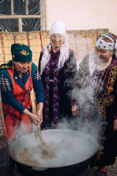 three women in traditional dress cooking food on an open fire pit with steam rising from it