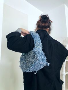 a woman in black jacket with blue crochet bag on her back, standing next to white wall