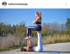 two women doing yoga poses on the ground near some water and trees, one woman is holding her leg up in the air