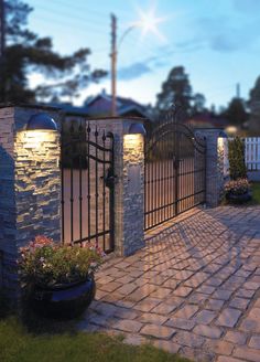 an iron gate is lit up at night in front of a brick driveway with potted plants