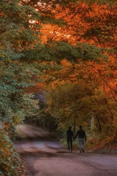 two people holding hands walking down a dirt road in front of trees with orange and yellow leaves