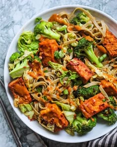 a white bowl filled with noodles, broccoli and tofu on top of a marble table