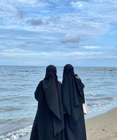 two women in burkas standing on the beach looking out at the water and sand