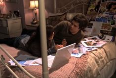 two people sitting on a bed in a bedroom with pictures and papers all over the floor