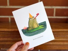 a hand holding up a card with a frog in a boat on the water wearing a hat