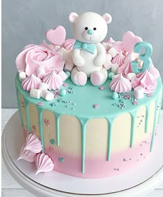 a birthday cake decorated with pink, blue and white icing has a teddy bear on top