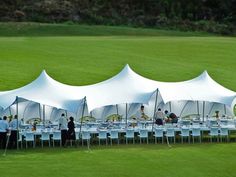 a large group of people sitting at tables under white tented tents on the grass