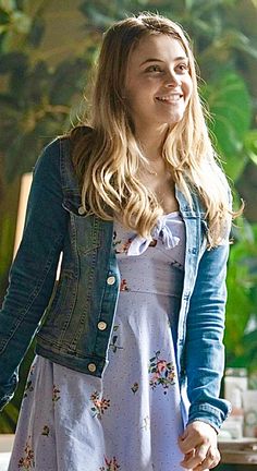 a woman in a dress and jean jacket smiles at the camera while holding her hand